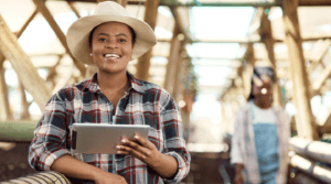 A Black woman in a hat and plaid shirt holds a mobile tablet in an agricultural space.