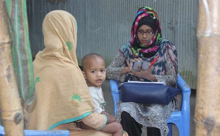 A woman interviews another woman using a tablet. The woman being interviewed is holding a small child.