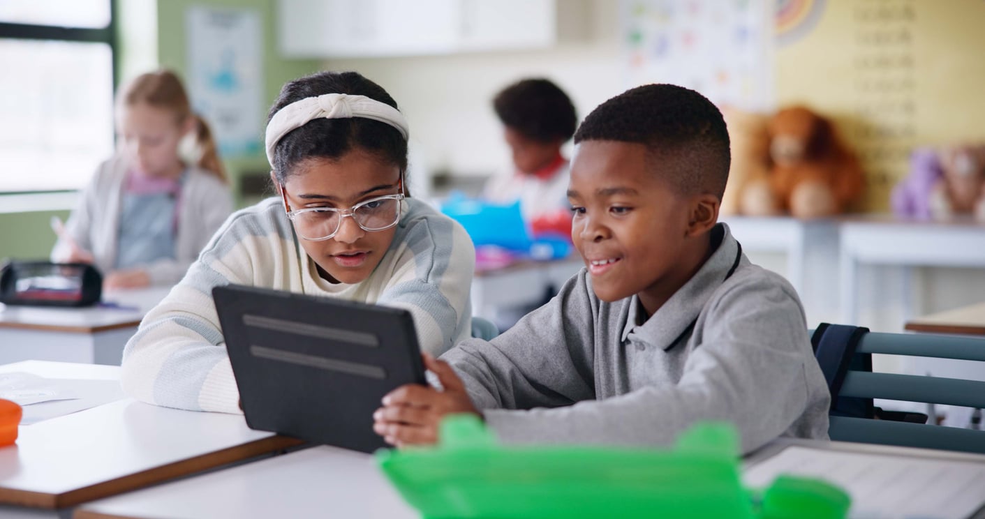 Two young children learn to read in a classroom by using a tablet.